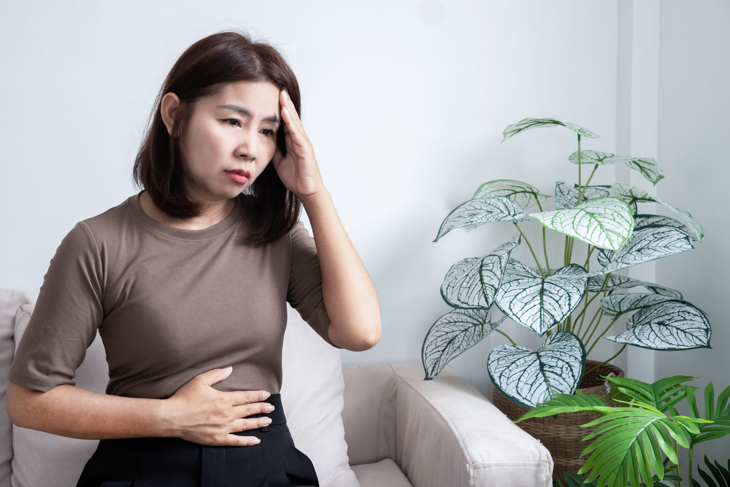 stressed Asian woman touching her head and stomach simultaneously. Concept of the gut-brain axis, indigestion caused by anxiety, mental health, and digestive problems in a bright living room